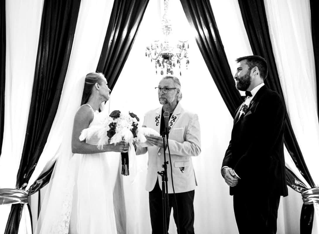 A bride and groom are holding hands while the officiant is giving them a toast.